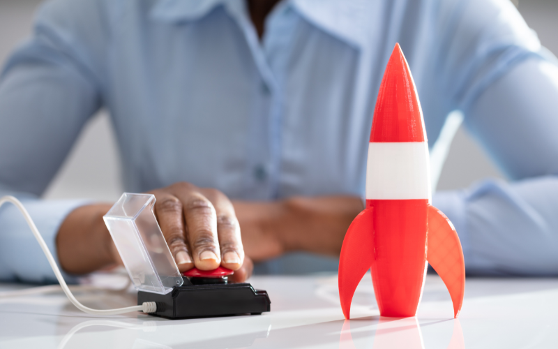 Person pressing a red button on a control box next to a red and white toy rocket on a table.